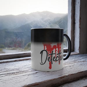 May include: A ceramic mug with a black-to-white gradient finish featuring the word Detective in black script lettering overlaid on a red dripping paint graphic. The mug has a black handle and is emitting a thin wisp of steam from the top opening.