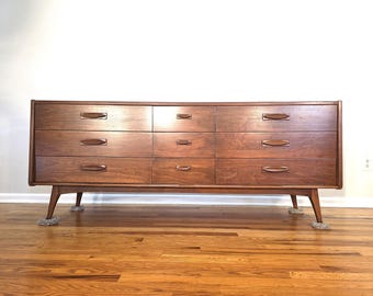 May include: A mid-century modern dresser with nine drawers. The dresser is made of wood with a warm brown finish. It has a rectangular shape with tapered legs and horizontal drawer pulls. The dresser is placed on a hardwood floor.