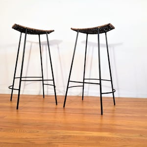 May include: Two black metal bar stools with woven brown seats. The stools have a simple, mid-century modern design.
