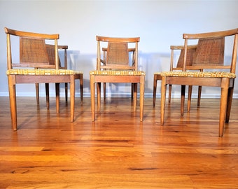 May include: Four wooden chairs with woven cane backs and yellow patterned upholstery. The chairs are arranged in a row on a hardwood floor.