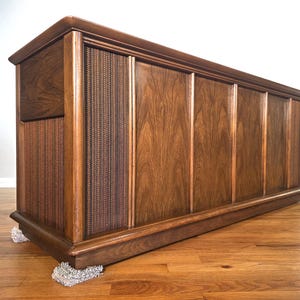 May include: A vintage wooden cabinet with a warm brown finish. The cabinet features a woven speaker cover on the left side and vertical wooden panels. It rests on small, light-colored pads on a hardwood floor.