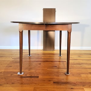 May include: A round wooden table with a brown finish and four legs. The table has a leaf that can be added to extend the table. The table is sitting on a hardwood floor.