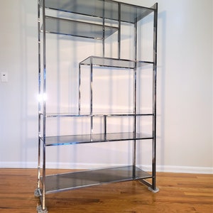 May include: A chrome and glass shelving unit with multiple shelves and a geometric design. The unit is standing on a wooden floor.
