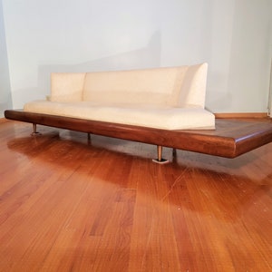 May include: A mid-century modern sofa with a cream-colored upholstered seat and backrest. The sofa sits on a dark wood base with metal legs. The design features a low profile and a minimalist aesthetic, suitable for a living room.