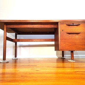 May include: A mid-century modern desk with a walnut finish. The desk has a rectangular top, a lower shelf, and a drawer unit on the right side. The desk is supported by four legs.
