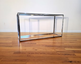 May include: A chrome console table with a rectangular top and a simple, open frame design. The table is made of polished chrome and has a sleek, modern look.