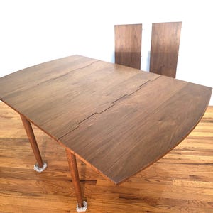 May include: A wooden, mid-century modern style drop-leaf table with a curved edge. The table is a medium brown color and has four legs. Two additional leaves are visible in the background. The table is approximately 48 inches long.