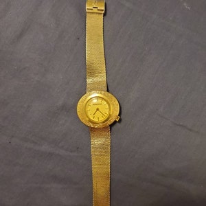 May include: A gold-toned wristwatch with a mesh band. The watch face is round with a textured bezel and gold hands. The band is a fine mesh, and the watch is fastened with a clasp. The watch is displayed on a dark gray surface.