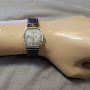 Könnte beinhalten: Eine silberne Armbanduhr mit weißem Zifferblatt und schwarzen römischen Ziffern als Stundenmarkierungen. Die Uhr hat ein braunes Lederarmband und befindet sich an einem Mannequin-Handgelenk.