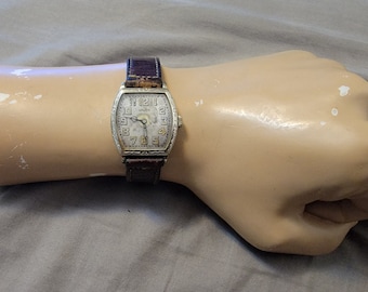 Può includere: Un orologio da polso vintage con cassa color argento a forma di botte e cinturino in pelle marrone scuro. Il quadrante presenta numeri arabi neri, un dettaglio circolare centrale e lancette sottili. La cassa in metallo è incisa.