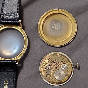 May include: A gold-toned wristwatch with a black leather band. The watch face is missing, and the back of the watch is open, revealing the inner workings of the watch movement.