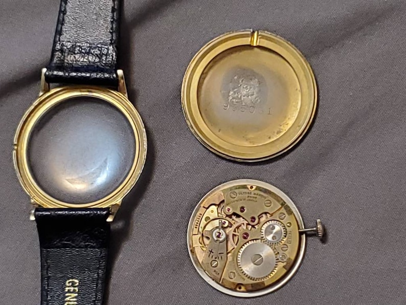 May include: A gold-toned wristwatch with a black leather band. The watch face is missing, and the back of the watch is open, revealing the inner workings of the watch movement.