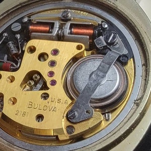 K&ouml;nnte beinhalten: Nahaufnahme des Innenlebens einer goldfarbenen Bulova-Uhr mit einer silbernen Batterie. Das Zifferblatt der Uhr ist mit "W.S.A. BULOVA 2181" beschriftet. Die R&uuml;ckseite der Uhr tr&auml;gt den Text "3306 ONLY GENUINE 400 CELL MARKED 218 STALL WITH BULOVA TOWARD CASE SIDE (218 TOWARD 0".
