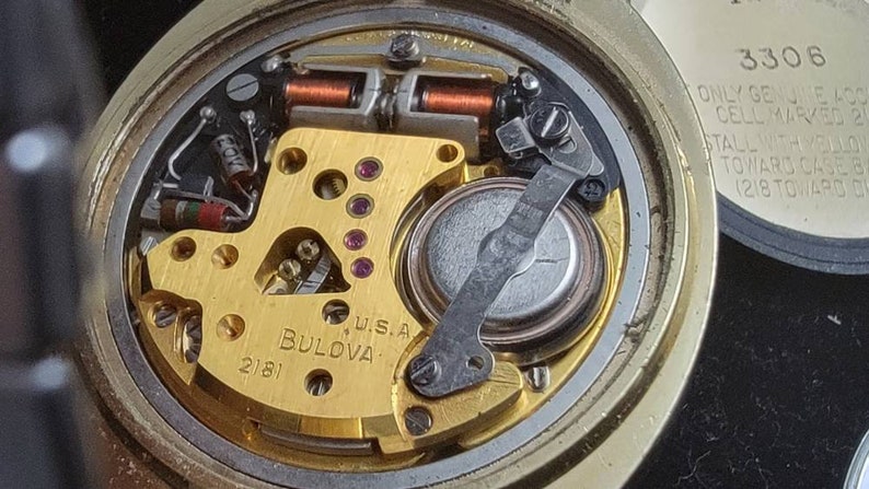 K&ouml;nnte beinhalten: Nahaufnahme des Innenlebens einer goldfarbenen Bulova-Uhr mit einer silbernen Batterie. Das Zifferblatt der Uhr ist mit "W.S.A. BULOVA 2181" beschriftet. Die R&uuml;ckseite der Uhr tr&auml;gt den Text "3306 ONLY GENUINE 400 CELL MARKED 218 STALL WITH BULOVA TOWARD CASE SIDE (218 TOWARD 0".