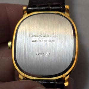May include: The back of a gold-tone watch with a brown leather band. The watch face is rectangular and has the text "Stainless Steel Back Waterresistant" and "7202/1" printed on it.