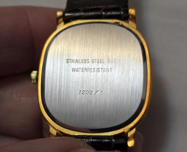 May include: The back of a gold-tone watch with a brown leather band. The watch face is rectangular and has the text "Stainless Steel Back Waterresistant" and "7202/1" printed on it.