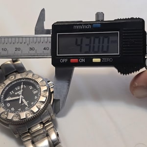 Puede incluir: Un reloj de pulsera plateado con una esfera negra y un bisel negro. El reloj se est&aacute; midiendo con un calibre digital, que lee 43,00 mm.