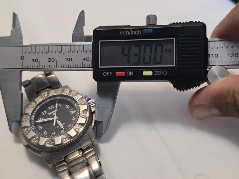 Puede incluir: Un reloj de pulsera plateado con una esfera negra y un bisel negro. El reloj se est&aacute; midiendo con un calibre digital, que lee 43,00 mm.