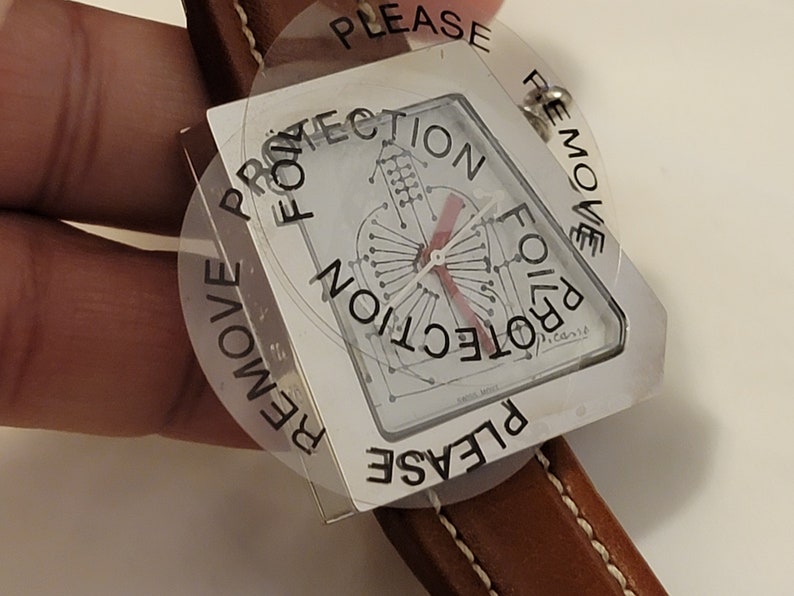 May include: A silver watch with a square face and a brown leather band. The watch face has a red and white design with the words "Protection Foil" printed around the edge. A clear plastic sticker with the words "Please Remove Protection Foil" is attached to the watch face.
