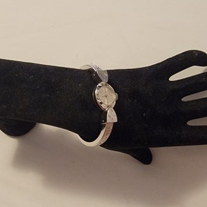 May include: A silver-toned bangle watch featuring a round white dial with silver hour markers and hands. The watch band is a rigid, hinged metal cuff with decorative engraved patterns on the sides, connecting to the central watch face.
