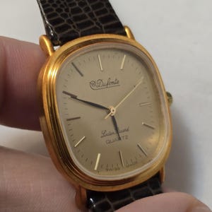 May include: A gold-tone wristwatch with a square face and a brown leather band. The watch face has a gold-tone dial with black hands and markers. The watch face also has the text "Duforte" and "Lucien Piccard QUARTZ" printed on it.