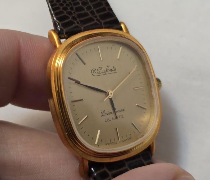 May include: A gold-tone wristwatch with a square face and a brown leather band. The watch face has a gold-tone dial with black hands and markers. The watch face also has the text "Duforte" and "Lucien Piccard QUARTZ" printed on it.