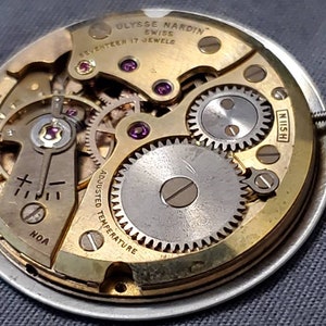 May include: Close-up of a gold-toned watch movement with the text "Ulysse Nardin Swiss Seventeen 17 Jewels" and "Adjusted Temperature" visible. The movement has a silver-toned winding stem and a series of gears.