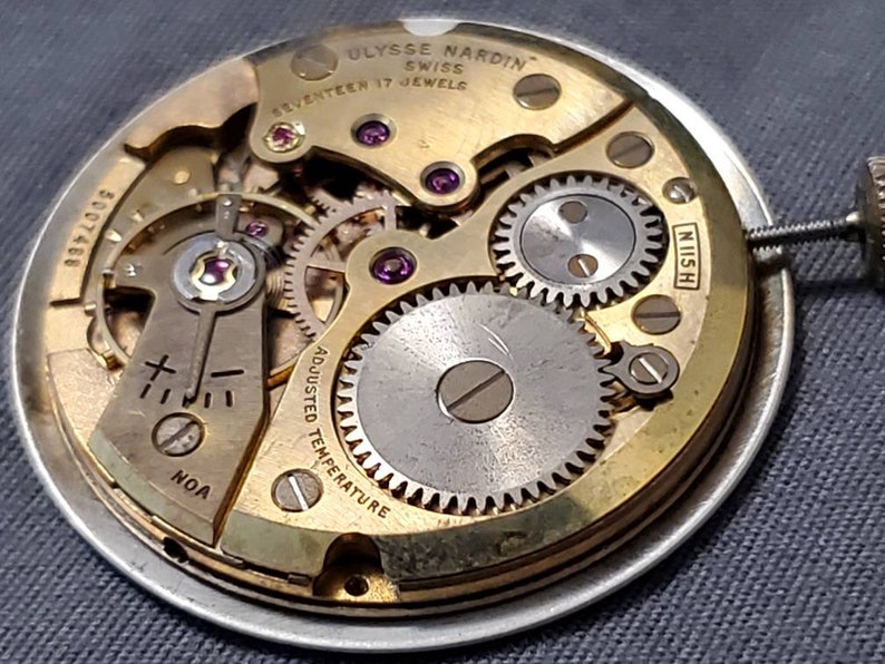 May include: Close-up of a gold-toned watch movement with the text "Ulysse Nardin Swiss Seventeen 17 Jewels" and "Adjusted Temperature" visible. The movement has a silver-toned winding stem and a series of gears.
