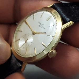 May include: A gold-tone wristwatch with a white face and black leather band. The watch face has black Roman numeral hour markers and a small seconds dial at the 6 o'clock position.