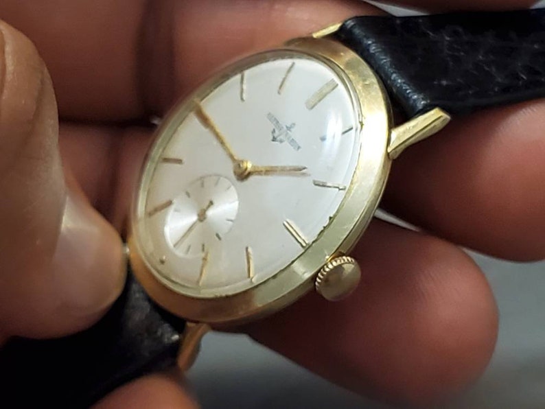 May include: A gold-tone wristwatch with a white face and black leather band. The watch face has black Roman numeral hour markers and a small seconds dial at the 6 o'clock position.