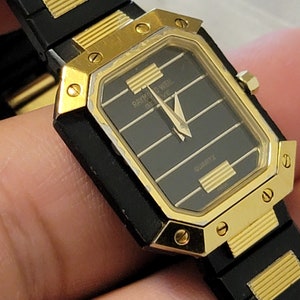 May include: A gold and black wristwatch with a square face and a black leather band. The watch face has gold Roman numeral hour markers and a black background. The watch face also has the text "Raymond Weil Geneve Quartz".