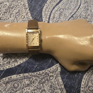 May include: A gold-toned rectangular watch with a brown leather band. The watch face has a white background with black Roman numeral hour markers and black hands.