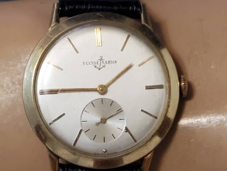 May include: A gold-tone wristwatch with a white face and black leather band. The watch face features gold-tone hands, hour markers, and a small sub-dial. The watch face also has the text "ULYSSE NARDIN" printed on it.