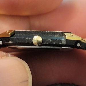 May include: A black and gold wristwatch with a square face and a black band. The watch has a gold-toned bezel and a gold-toned crown.