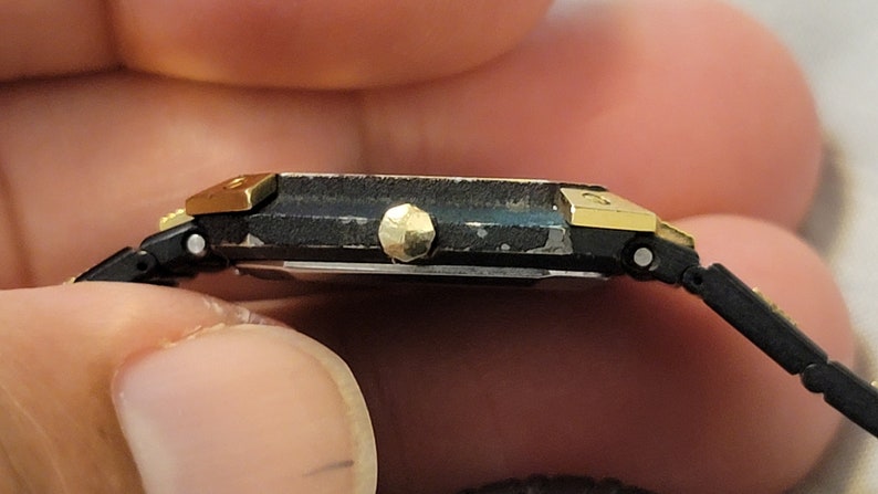 May include: A black and gold wristwatch with a square face and a black band. The watch has a gold-toned bezel and a gold-toned crown.