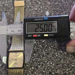 May include: A gold-tone watch with a square face and a two-tone metal band. The watch face is a light gold color with black Roman numeral hour markers and black hands. The watch band is a combination of gold and silver metal links. The watch is being measured with a digital caliper, which reads 25.00 mm.