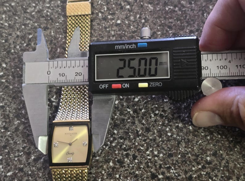 May include: A gold-tone watch with a square face and a two-tone metal band. The watch face is a light gold color with black Roman numeral hour markers and black hands. The watch band is a combination of gold and silver metal links. The watch is being measured with a digital caliper, which reads 25.00 mm.