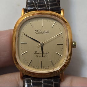 May include: A gold-tone wristwatch with a square face and black leather band. The watch face has a white background with black hands and black markings. The watch face also has the text "Duforte" and "Lucien Piccard QUARTZ" printed on it.
