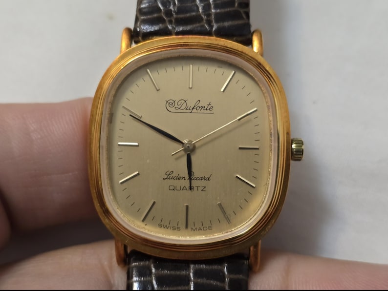 May include: A gold-tone wristwatch with a square face and black leather band. The watch face has a white background with black hands and black markings. The watch face also has the text "Duforte" and "Lucien Piccard QUARTZ" printed on it.