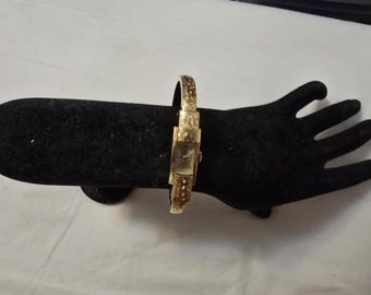 May include: A gold-toned wristwatch with a square face and a textured band. The watch is on a black display stand.
