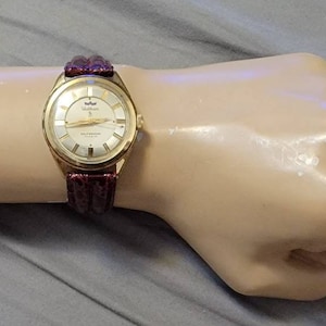 Puede incluir: Un reloj de pulsera dorado con una esfera blanca y una correa de cuero burdeos. La esfera del reloj tiene una ventana de fecha y las palabras "Sea-Commander" impresas en ella.