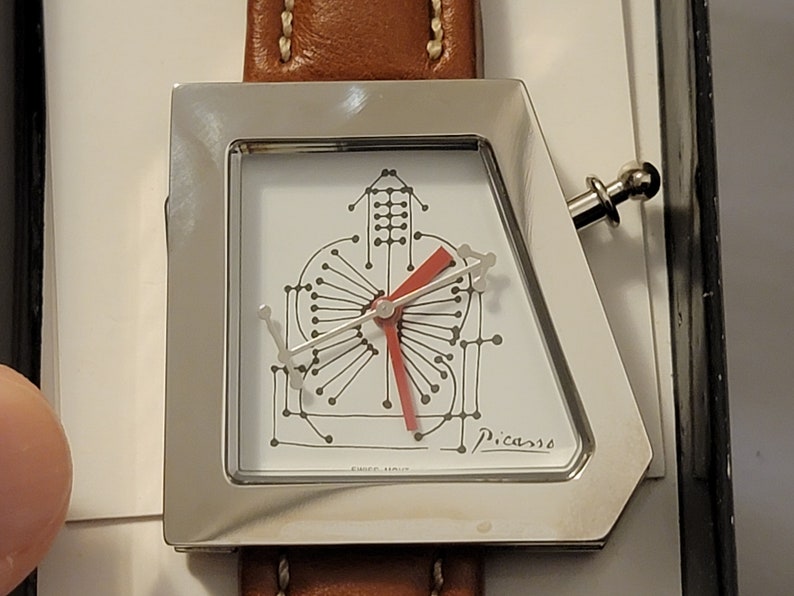 May include: A silver watch with a white face and a red second hand. The watch face has a unique design with a black and white pattern and the word "Picasso" printed on the bottom. The watch is attached to a brown leather band.