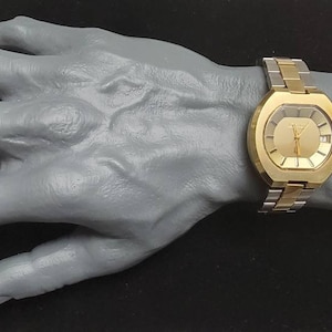 K&ouml;nnte beinhalten: Eine goldfarbene Armbanduhr mit quadratischem Zifferblatt und einem zweifarbigen Metallband. Die Uhr hat ein goldenes Zifferblatt mit schwarzen Zeigern und Markierungen.