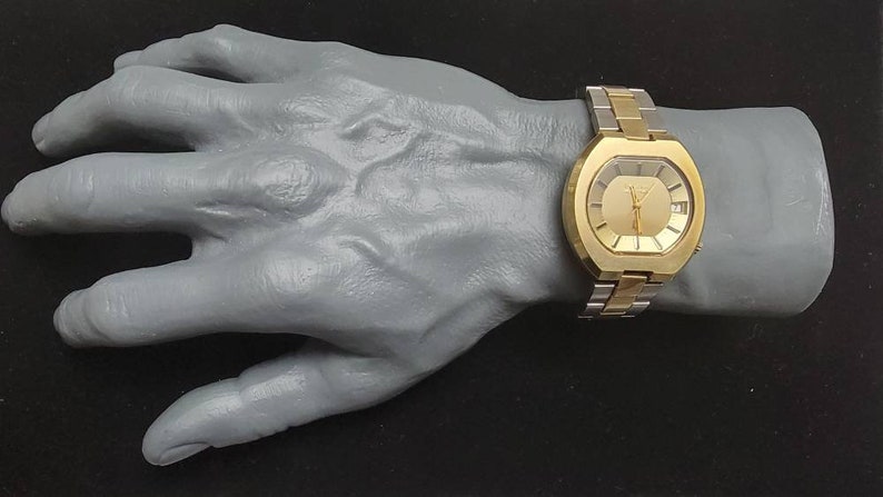 K&ouml;nnte beinhalten: Eine goldfarbene Armbanduhr mit quadratischem Zifferblatt und einem zweifarbigen Metallband. Die Uhr hat ein goldenes Zifferblatt mit schwarzen Zeigern und Markierungen.