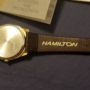May include: A gold-toned Hamilton wristwatch with a brown leather strap. The watch face is visible, along with the engraved text "HAMILTON" on the strap. The watch back has engraved text including "CHARLES F PETERS" and "STAINLESS STEEL BACK".
