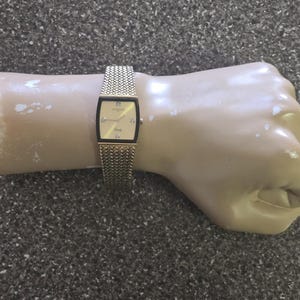 May include: A gold-tone watch with a square face and a chain link band. The watch face has a white background with black Roman numeral hour markers and black hands.