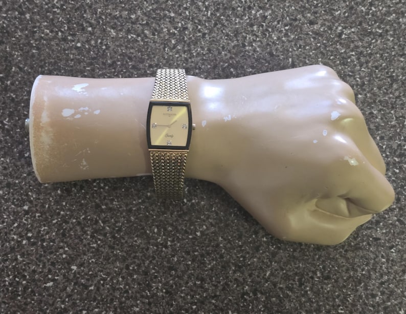 May include: A gold-tone watch with a square face and a chain link band. The watch face has a white background with black Roman numeral hour markers and black hands.