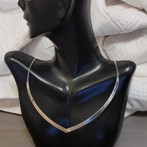 May include: A silver-colored choker necklace with a V-shaped design. The necklace is displayed on a black mannequin bust against a white fabric background. The choker is a simple, elegant piece of jewelry.