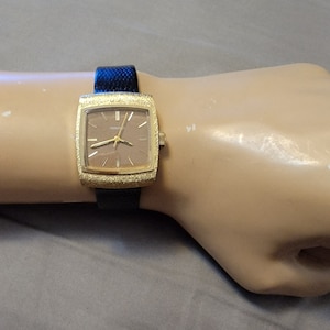 May include: A gold-toned square wristwatch with a brown face and black band. The watch is on a mannequin wrist.