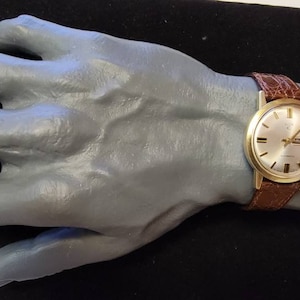 May include: A gold-toned wristwatch with a brown leather band is displayed on a gray hand-shaped form. The watch has a light-colored face with gold markers and hands. The band has a crocodile pattern.